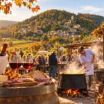 Veduta autunnale dell’Oltrepò Pavese con borgo collinare, calici e bottiglie di vino e scena di festa gastronomica all’aperto.